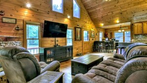 A cozy log cabin interior with a large TV, leather sofas, a fireplace, and wooden walls.