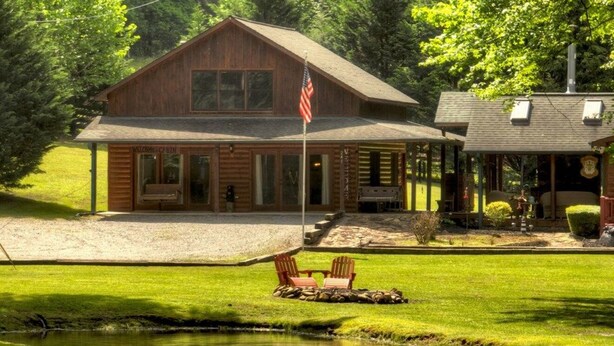 A cabin with a pond, two Adirondack chairs, and a flag.
