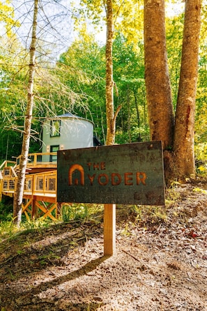 A rustic wooden sign with the text 'THE AYODER' in a forested area with a small cabin and wooden deck. - NEW Romantic Silo Cabin | Outdoor Shower | Hot Tub (Beattyville)