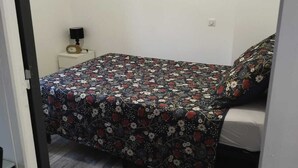 Ein kleines Zimmer mit einem Einzelbett, einer Bettdecke mit Blumenmuster, einem Nachttisch mit Lampe und einem Holzboden.