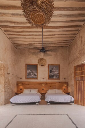 Une chambre avec une tête de lit en bois, deux lits, un luminaire suspendu en fibres tressées et deux tableaux encadrés accrochés au mur.