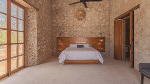 Quarto com parede de pedra, cama de madeira, ventilador de teto e uma janela grande.