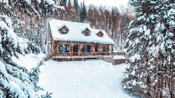 Una cabaña de troncos con techo cubierto de nieve, rodeada de árboles cargados de nieve.