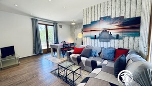 A modern living room with a sofa, a coffee table, and a TV on a stand. There is a dining area with a table and chairs. The room has a large window with curtains. - La Ceriseraie - TRAUSSE (TRAUSSE)