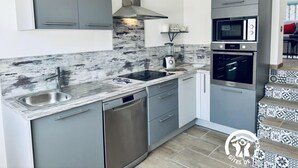 A modern kitchen with grey cabinets, a stainless steel dishwasher, a built-in oven, and a marble backsplash. - La Ceriseraie - TRAUSSE (TRAUSSE)