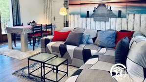 A modern living room with a sectional sofa, a dining table, and a large painting of a sunset over a pier. - La Ceriseraie - TRAUSSE (TRAUSSE)