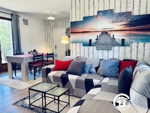 Ein modernes Wohnzimmer mit einer Eckcouch, einem Esstisch und einem großen Gemälde, das einen Sonnenuntergang über einem Steg zeigt.