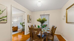 A dining area with a wooden table set for four, surrounded by chairs. There are two framed artworks on the walls, a potted plant, and a hanging light fixture.