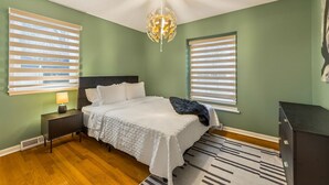 A bedroom with a bed, nightstands, a lamp, a ceiling light, and two windows with blinds.