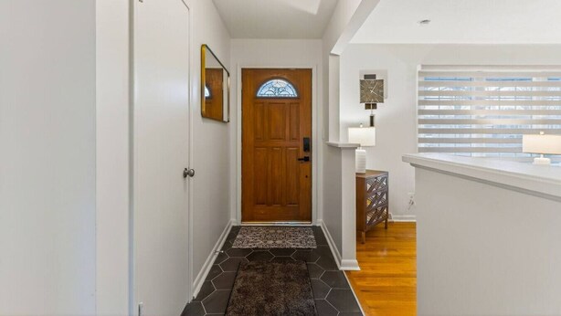 A hallway with a wooden door, a small rug, a wall-mounted clock, and a window with blinds.
