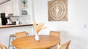 En matplats med ett runt träbord, sex stolar, en vävd matta, en väggmonterad lampa och ett kök i bakgrunden.