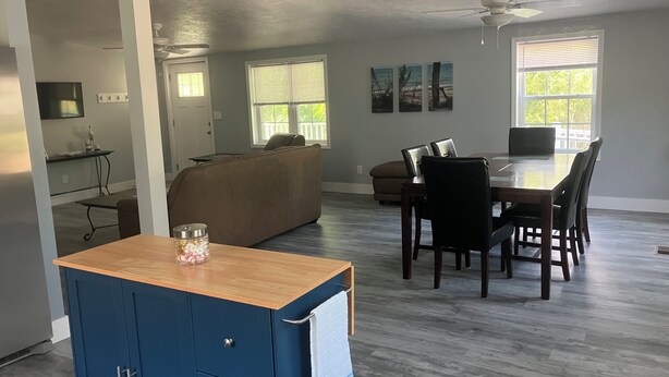 A spacious living area with a dining table, a sofa, and a kitchen area with a blue cabinet. - Perfect 3-bedroom mobile home with AC in charming Albion. Mobility-accessible. (Albion)