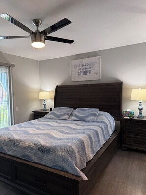 Une chambre à coucher avec un lit, deux tables de chevet avec des lampes, un ventilateur de plafond, une carte du monde décorative et une fenêtre avec des stores.