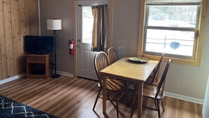 Una habitación con piso de madera, una mesa de comedor con sillas, un televisor sobre un mueble, una lámpara y una ventana que da a un área cercada.