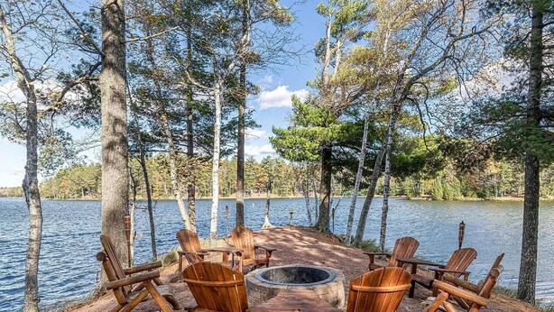 Un hoyo para fuego circular rodeado de sillas Adirondack de madera, sobre una zona de grava, con un lago y árboles al fondo.