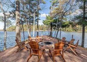 Ein runder Feuerkorb, umgeben von Holz-Adirondack-Stühlen, auf einer Schotterfläche mit einem See und Bäumen im Hintergrund.
