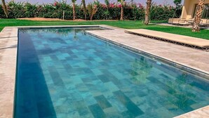 Une piscine rectangulaire à l’eau claire, entourée d’une terrasse carrelée et d’une zone herbeuse agrémentée de palmiers et d’arbustes.