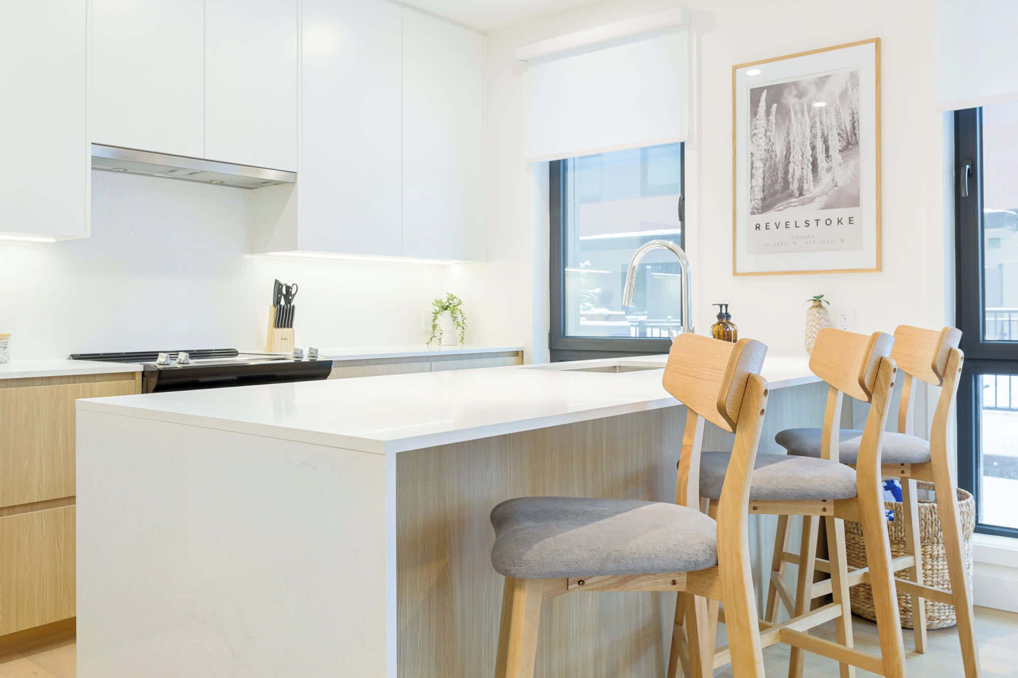 A modern kitchen with a central island, wooden bar stools, and a large window providing natural light.