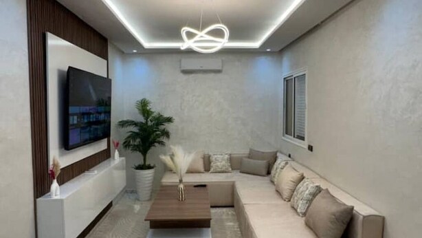 Una sala moderna con un televisor de pantalla plana montado en la pared, una mesa de centro de madera, un sofá beige con cojines y una planta en maceta.