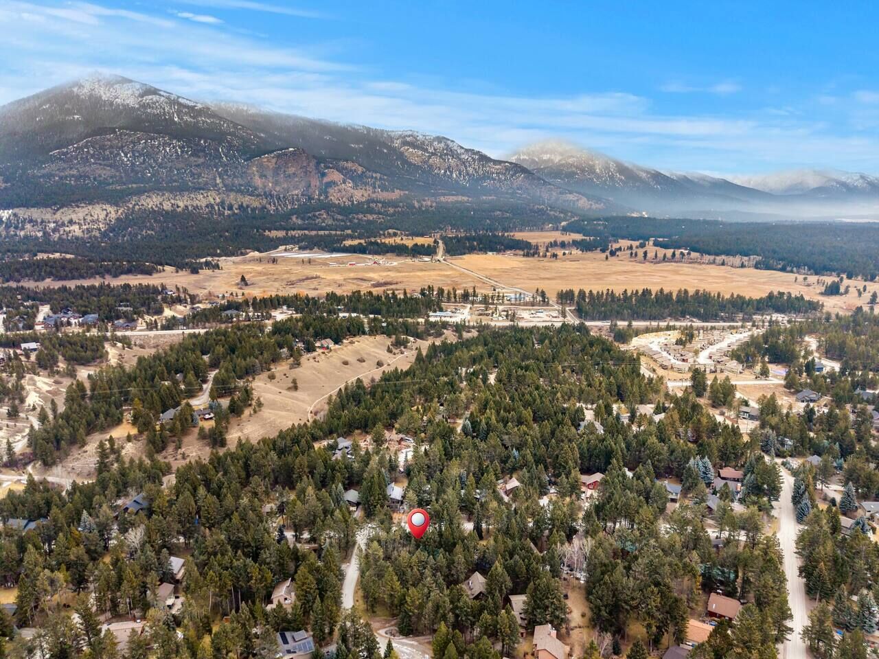 Vue aérienne d’un quartier résidentiel, avec des maisons entourées de forêts denses, un ciel dégagé et des montagnes en arrière-plan.