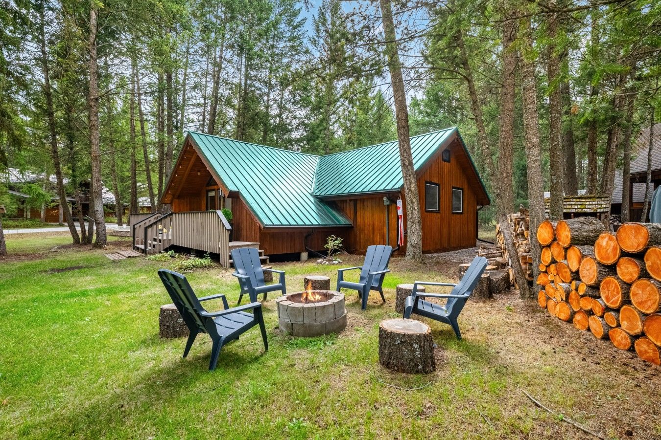 A cabin with a green roof, surrounded by trees, and a fire pit with chairs.
