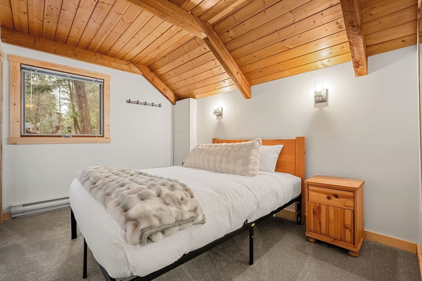 A bedroom with a wooden ceiling, a large bed with a fluffy blanket, a nightstand, and a window with a view of trees.