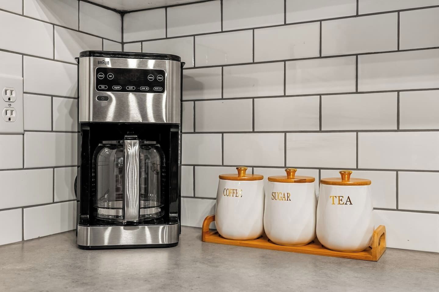 A coffee maker with a digital display and a coffee bean grinder. There are three white containers with wooden lids labeled "Coffee," "Sugar," and "Tea."