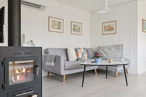 A living room with a wood-burning stove, a grey sofa, a coffee table, and framed artwork on the walls. - Bright holiday home with sunny terrace (Væggerløse)