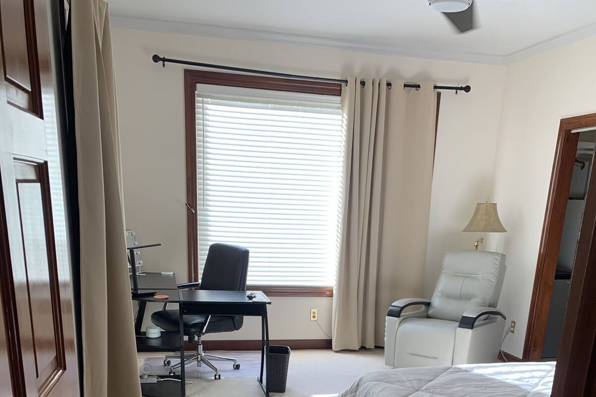 A hotel room with a bed, a desk with a chair, a small table, a lamp, and a window with curtains.