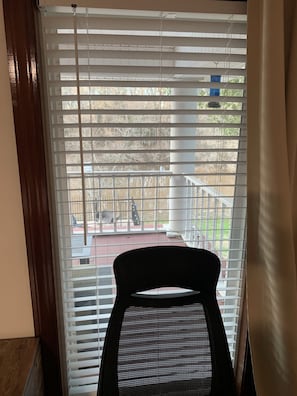 A room with a view through a window with blinds, a black chair, and a railing outside.