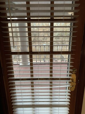 A wooden door with white blinds partially open, revealing a view of a red-tiled patio and a white column outside.