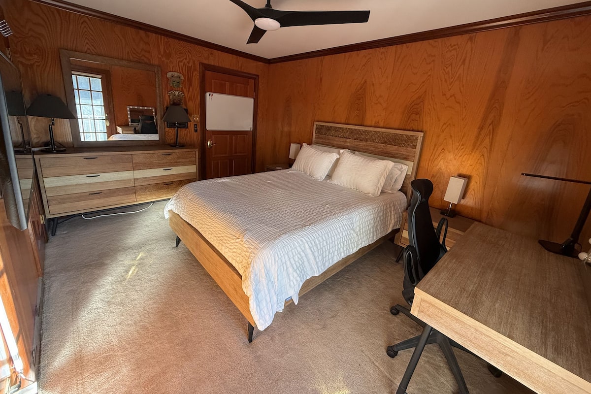 A bedroom with a wooden headboard, a bed with white bedding, a desk with a chair, a ceiling fan, and a dresser with a mirror.