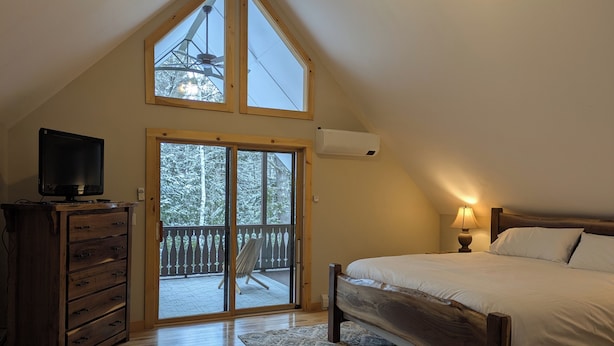 Camera da letto con soffitto spiovente, un letto, una TV su una cassettiera in legno e un balcone con vista sugli alberi.