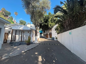 A narrow street with a white building on the left, a tree-lined path, and a white wall with a green sign on the right.