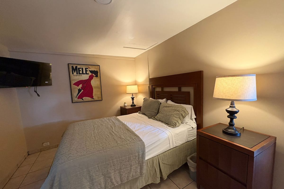 A bedroom with a bed, a nightstand, a lamp, a TV mounted on the wall, and a poster.