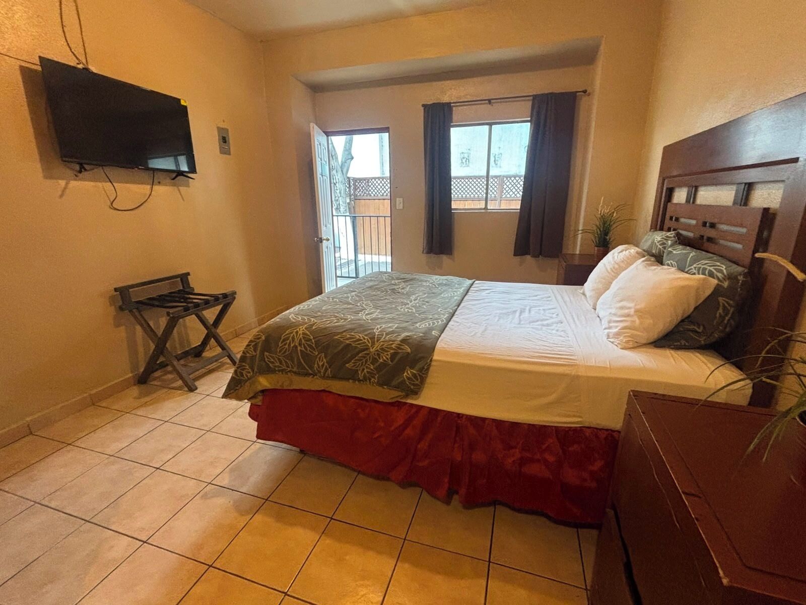 Un dormitorio con cama, televisión montada en la pared, una mesita y una ventana con cortinas.