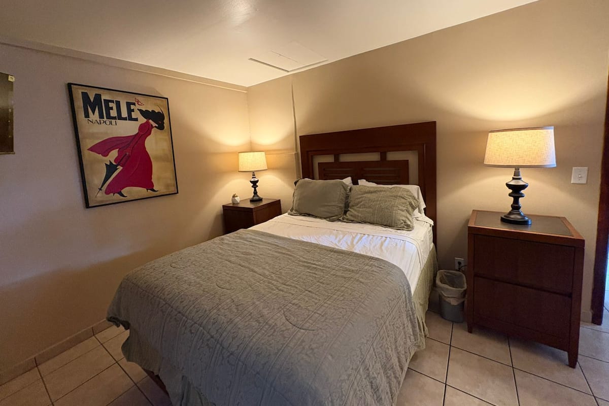 A hotel room with a bed, two bedside tables, a nightstand, a trash bin, and a poster on the wall.