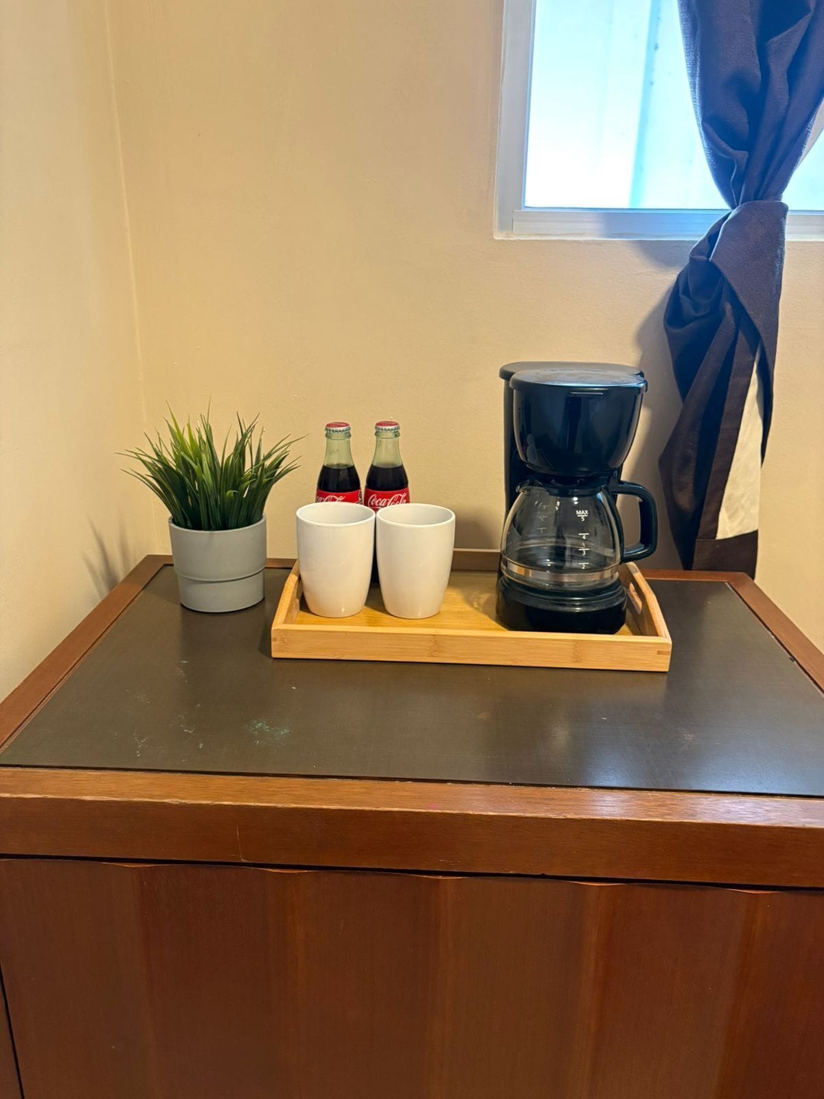 Una mesa de madera con una cafetera, dos tazas blancas, dos botellas de Coca-Cola y una planta en maceta.