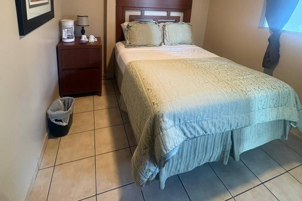 A hotel room with a bed, a nightstand, a framed picture, a window with curtains, and a tiled floor.