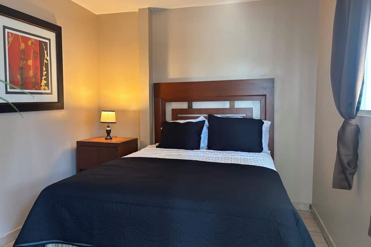 A hotel room with a bed, a nightstand, a lamp, a framed picture on the wall, and a window with curtains.