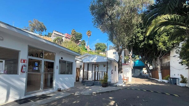 A street view with a white building with a large window and a fire extinguisher sign, a small white building with a door and steps, and a tree with a curved trunk. - Motel del Bosque (Tijuana)
