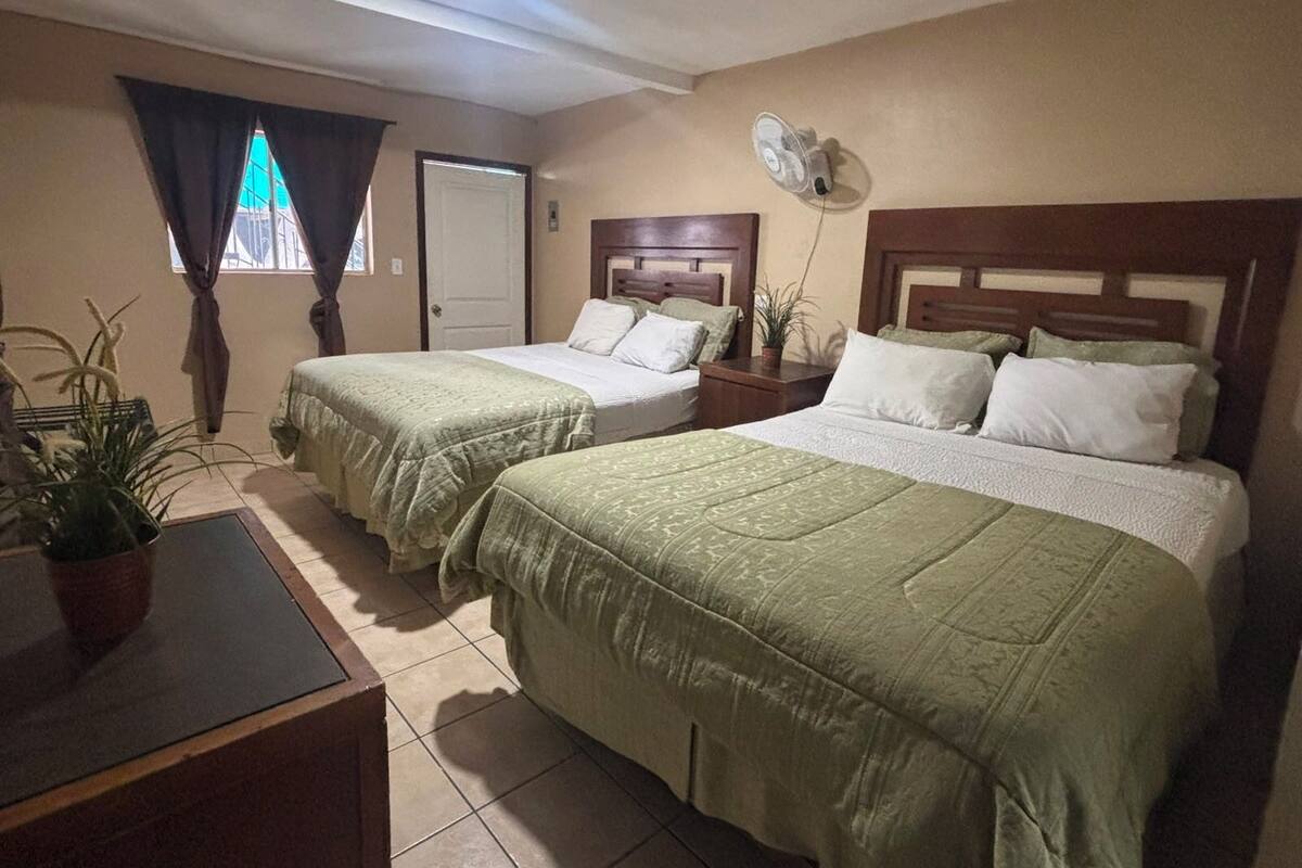 A hotel room with two beds, a bedside table, a fan, and a window with curtains.