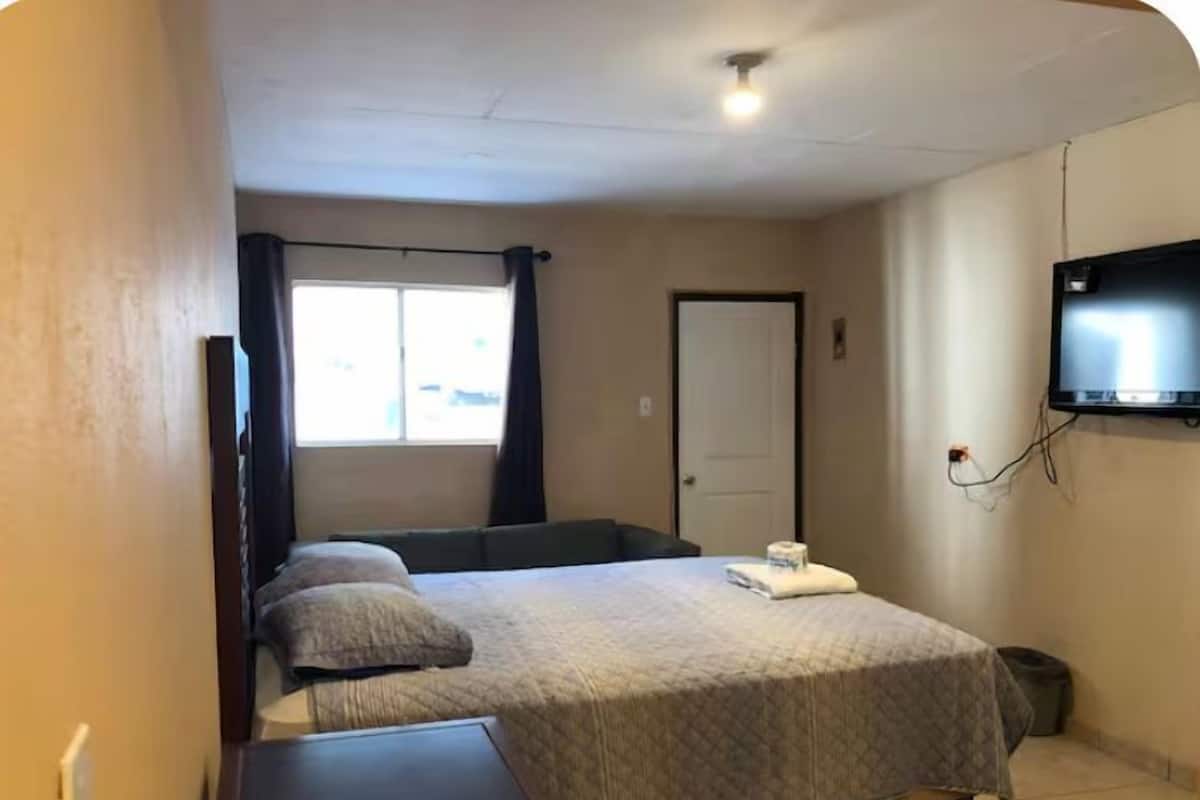 A bedroom with a bed, a desk, a television mounted on the wall, and a window with curtains.