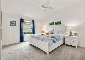 Ein Schlafzimmer mit einem weißen Bett, einem Nachttisch, einem Deckenventilator und einem Fenster mit blauen Vorhängen.
