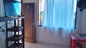 A room with a bunk bed, a wooden door, a window with white curtains, a red table, and a ceiling fan. - Hotel Volga (Puerto Lopez)