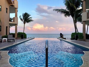 Ein Pool mit Blick auf den Ozean bei Sonnenuntergang, umgeben von Palmen und einem Gebäude mit Balkonen.