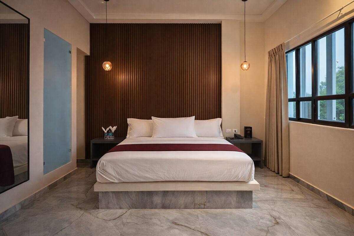 Une chambre d’hôtel moderne avec un grand lit, un mur lambrissé en bois, deux lampes suspendues, une table de chevet et une fenêtre avec des rideaux.