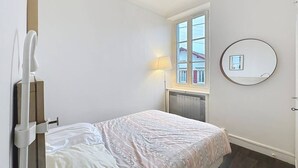 Een slaapkamer met een bed, een raam, een radiator, een lamp en een ronde spiegel aan de muur.