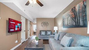 A living room with a flat-screen TV mounted on the wall, a grey sofa, a coffee table with a plant, and a painting of trees on the wall. - Hopes Place Near Ft Campbell (Clarksville)