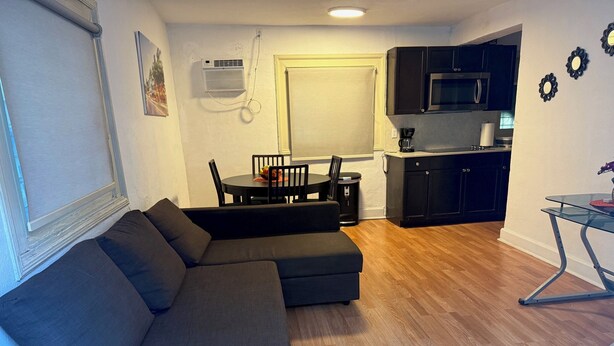 A living room with a sectional sofa, a dining table with chairs, a kitchen area with cabinets and a microwave, and a small table with a vase.
